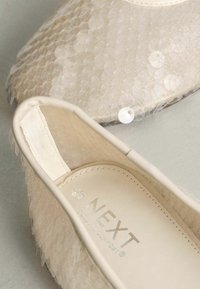 Bēšas baleta kurpes ar fakturētu, zvīņas rakstu, caurspīdīgu augšdaļu, mīkstu materiālu un iespiestu "NEXT" zīmolu iekšpusē.