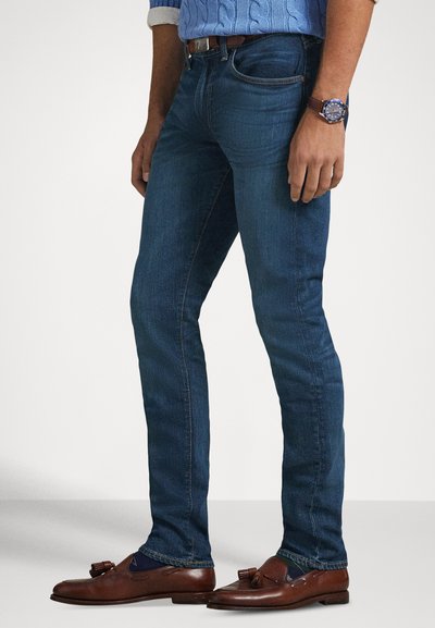 Jeans de ganga azul escuro com corte slim, apresentando costuras contrastantes e cintura média, combinados com mocassins castanhos.