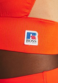 Un tissu côtelé orange présente un écusson brodé blanc et bleu avec le logo "BOSS HUGO BOSS" sur un vêtement de sport.