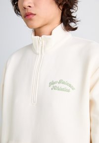 Pull à col montant crème en polaire, avec une fermeture éclair quart, présentant une texture douce et un logo "New Balance Athletics" brodé en vert sur la poitrine.