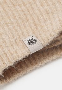 Beige ribbed knit fabric with a soft texture, featuring a white woven label with a black emblem attached to the edge.