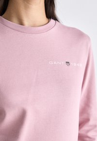 Gros plan d’une personne portant un sweat-shirt rose clair avec « GANT 1949 » et un logo en forme de bouclier sur la poitrine, sur un fond uni.