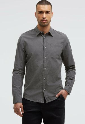 Mann steht frontal, trägt ein langärmliges graues Hemd mit Knöpfen und Brusttasche sowie schwarze Hose, rechte Hand in der Hosentasche.