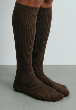 Jambes portant des chaussettes hautes côtelées marron foncé, debout sur un sol gris devant un fond clair uni.
