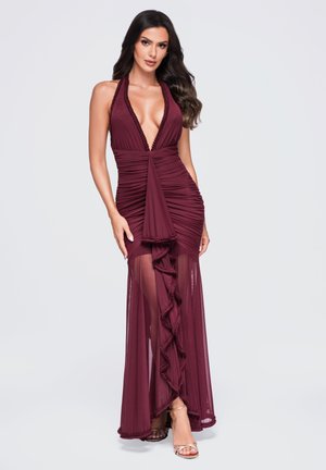 Frau in einem ärmellosen bordeauxfarbenen Kleid mit tiefem V-Ausschnitt und transparentem Rock-Volant-Detail, posiert vor einem schlichten hellen Hintergrund.