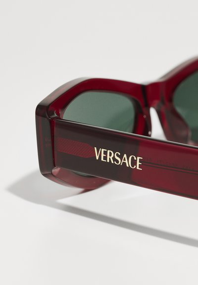Raudonos akrilinės saulės akinių su šešiakampiu pavidalu, tamsiai žali objektyvai ir auksinis "Versace" logotipas šone, su tekstūruota detalizacija ant šakės.