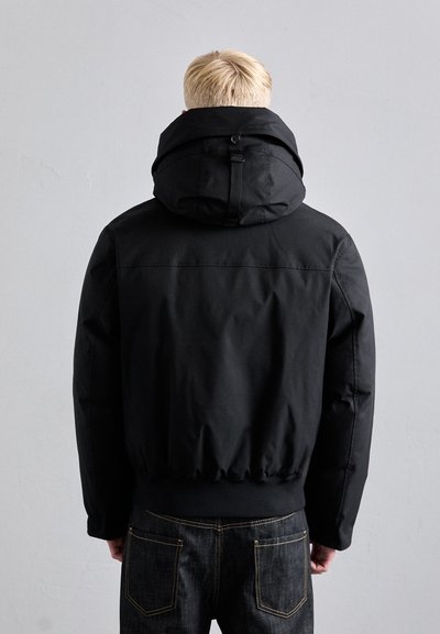 Black puffer jacket with a hood, elastic cuffs, and a cinched hem. Smooth texture and minimalistic design. Paired with dark jeans.