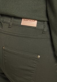 Olivgröna byxor med en formad bakficka, som har en NAF NAF-logotag. Den släta texturen och subtila sömsdetaljer framhäver designen.