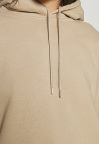 Beżowa bluza z kapturem, wykonana z fakturowanej tkaniny, z kapturem na sznurku i sznurkami z metalowymi końcówkami. Prosty, minimalistyczny design.
