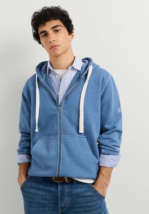 Junger Mann mit lockigem Haar, der einen blauen Kapuzenpullover mit Reißverschluss über einem gestreiften Shirt und Jeans trägt, steht mit den Händen in den Taschen vor einfarbigem Hintergrund.