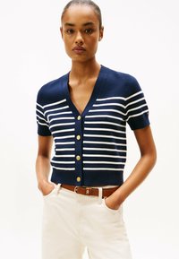 Cardigan navy a maniche corte con strisce bianche, design a V, con bottoni dorati e silhouette aderente. Abbinato a jeans chiari.