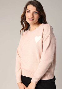 Pull en maille rose avec un col rond et des poignets côtelés. Présente un petit cœur blanc sur la poitrine gauche. Texture douce, coupe décontractée.