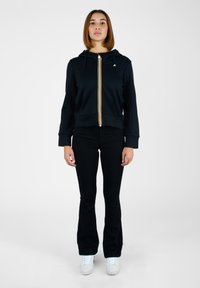 Felpa con zip di colore navy con righe gialle e bianche, con cappuccio regolabile, indossata su pantaloni neri svasati e sneakers bianche.