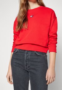 Rode sweater met lange mouwen en ribgebreide boorden, voorzien van een klein geborduurd logo. Gepaarde met zwarte high-waisted jeans.