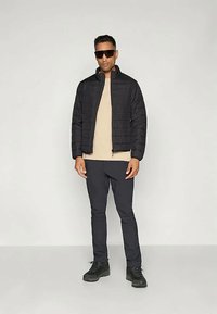 Juodas izoliuotas švarkas su užtrauktuku priekyje, vertikaliu qiltu dizainu, derinamas su smėlio spalvos marškinėliais ir tamsiomis kelnėmis, užbaigtas juodais batais.