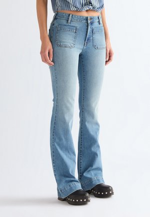 Woman wearing light blue flared jeans with front patch pockets and black studded shoes against a plain background.