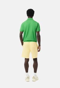 Camisa polo verde, pantalones cortos amarillos, calcetines blancos hasta el tobillo y zapatillas beige. La polo es de manga corta y tiene cuello; los pantalones cortos son hasta la rodilla.