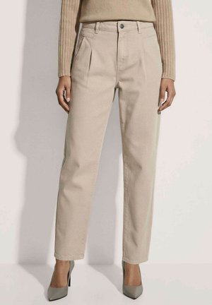 Vrouw draagt beige plissé broek met hoge taille, gecombineerd met grijze hoge hakken en een beige geribde trui.