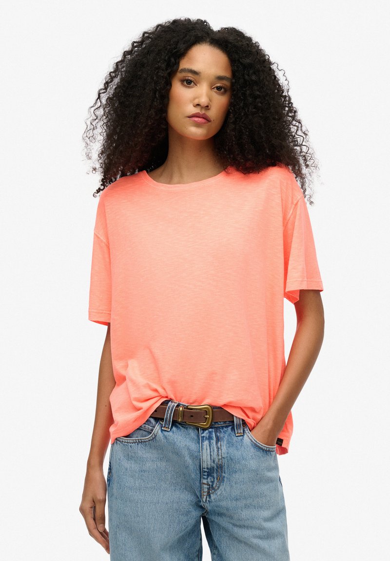 Camiseta de cuello redondo en color coral, fabricada en tejido ligero, de corte relajado y con mangas cortas. Combinada con jeans de mezclilla claros de cintura alta y un cinturón marrón.