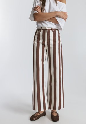 Personne portant un pantalon taille haute à jambes larges avec des rayures verticales marron et blanches, un top blanc aux épaules dénudées et des chaussures plates tressées marron.