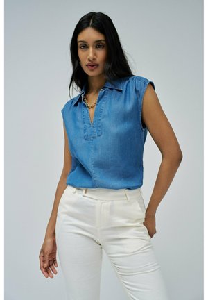 Blusa de mezclilla sin mangas en azul claro con cuello tipo notch y sutiles detalles de pliegues, combinada con pantalones sastreros de color crema.