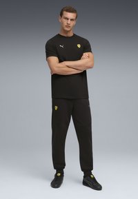 Czarna sportowa koszulka i spodnie z żółtym logo Ferrari. Materiał wydaje się miękki, o gładkiej fakturze. Czarne sneakersy dopełniają strój.