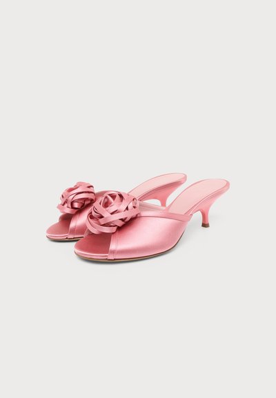 Mules à talons roses en satin à bout ouvert avec des rosettes décoratives en ruban sur le dessus, sur fond blanc.