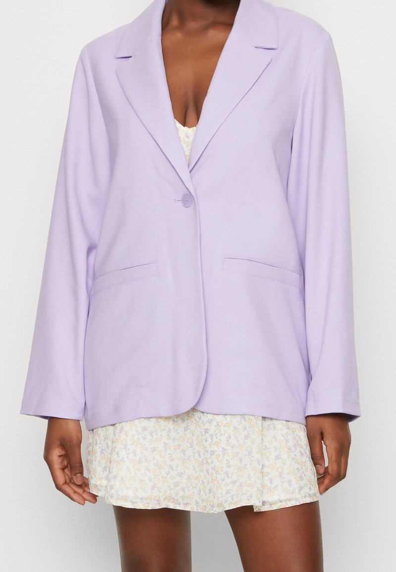 Femme portant un blazer lilas clair à un seul bouton sur une mini robe blanche fleurie, sur un fond uni.