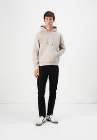 Helle graue Kapuzenjacke mit einer Fronttasche und einer Kordelzugkapuze, kombiniert mit schwarzen Jeans und weißen Sneakers mit schwarzen Akzenten.