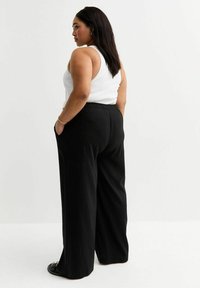 Pantaloni neri a gamba larga con una vestibilità rilassata e cintura elastica, abbinati a una maglietta bianca senza maniche. Il modello è in piedi con una leggera inclinazione.