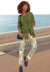 Grüne Strickjacke über einem strukturierten grünen Oberteil, kombiniert mit Blumenmuster-Hosen, die pinke und orangefarbene Blumen zeigen. Marineblaue Loafers und eine Umhängetasche.