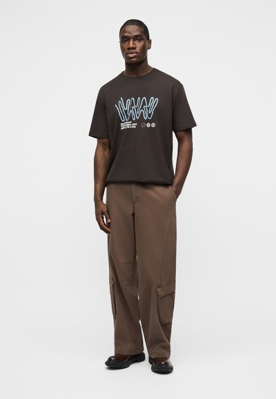 Άνδρας στέκεται με το πρόσωπο προς τα εμπρός, φοράει σκούρο καφέ μπλουζάκι με γραφικά, φαρδιά καφέ παντελόνια cargo και μαύρα παπούτσια, με τα χέρια χαλαρά δίπλα στα πλευρά.