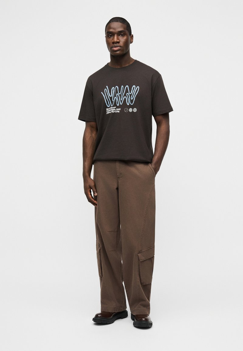 Man stands facing forward wearing dark brown graphic t-shirt, loose brown cargo pants, and black shoes, hands relaxed by sides.