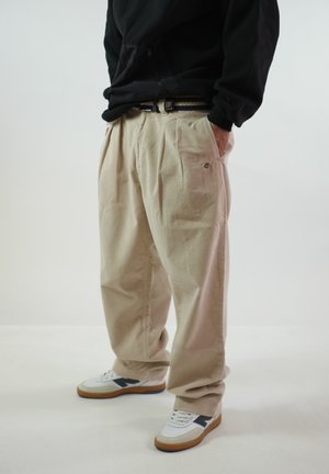 Persoon die beige geplooide broek draagt, zwarte longsleeve, witte sneakers met zwarte en bruine accenten, handen in de zakken, staand op een witte vloer.