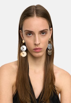 Młoda kobieta z prostymi brązowymi włosami nosi duże, wielodisowe złote i białe kolczyki wiszące oraz czarny bezrękawnik na jednolitym tle.