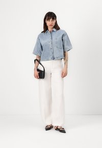 Giacca di jeans corta con tasca sul petto, indossata sopra pantaloni bianchi in lino. Borsa a spalla nera con manico e scarpe nere basse con fiocchi.
