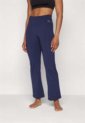 Kobieta ubrana w granatowe legginsy do jogi z wysokim stanem marki Puma i czarny krótki top, stojąca boso na tle jednolitej białej ściany.