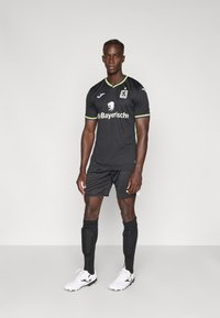 Melns futbola tērps ar īsām piedurknēm, kurā redzams lauva logo un sponsora teksts, izcelts ar zaļām, dzeltenām un zilām svītrām apkaklē un piedurknēs.