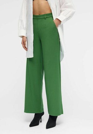 Pantalon vert taille haute et jambe large, associé à des bottines à talons noires à bouts pointus et une longue chemise blanche boutonnée.