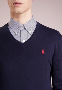 Marineblauer V-Ausschnitt-Pullover aus weichem Strickmaterial, mit einem roten gestickten Logo auf der linken Brust, kombiniert mit einem gestreiften Shirt.