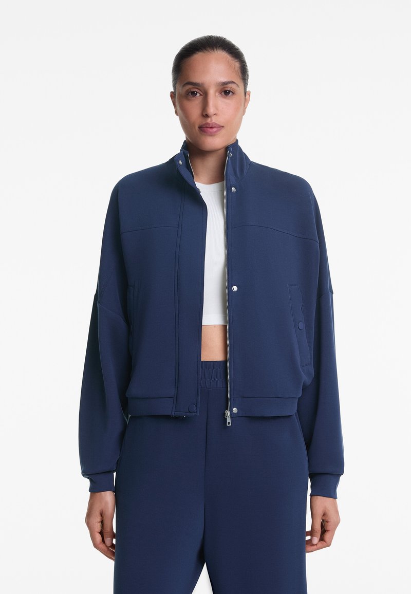 Dunkelblaue cropped Jacke mit hohem Kragen, Druckknöpfen und seitlichen Taschen, aus einem glatten, dehnbaren Stoff.