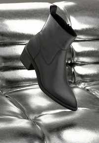 Botă de gleznă din piele neagră cu toc mic, așezată pe o suprafață strălucitoare, metalică, de culoare argintie, evidențiind textura netedă și designul elegant.