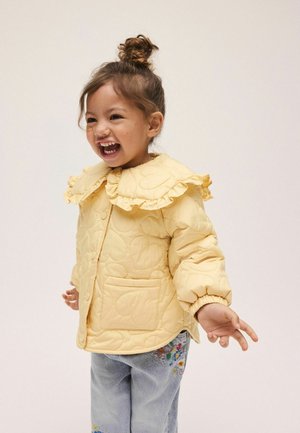 Gul quiltet jakke med rynket krave, med knaplukning foran og en sidelomme. Model iført blå jeans med blomsterbroderi.