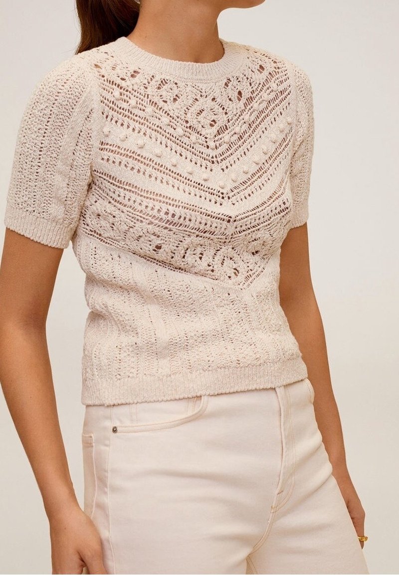 Žena oblečená v krémovém pleteném topu s krátkým rukávem, který má složité krajkové vzory a detaily s bambulkami, doplněném o krémové kalhoty s vysokým pasem.