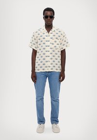 Korte mouw, crèmekleurig shirt met blauwe oogmotieven, gecombineerd met lichtblauwe jeans en witte sneakers. Model draagt een donkere zonnebril.