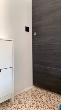 Weißer Schrank mit schwarzem Griff, graue Holzwand, runde Wandlampe und gemusterter Terrazzo-Boden mit roten, beigen und weißen Farbtönen.
