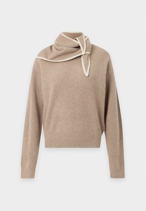 Femme debout avec de longs cheveux bouclés portant un pull beige, une jupe courte brun foncé avec un ourlet à volants blancs et des sandales noires à talons hauts.