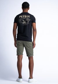 Musta puuvillapaita, jossa on kultapainatus, jossa lukee "PETROL" ja rantalainen maisema. Yhdistetty oliivinvihreisiin shortseihin ja valkoisiin tennareihin.