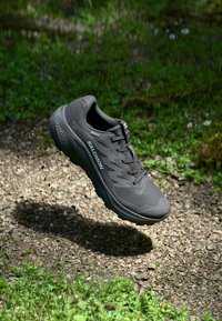 Black running shoe with textured upper, cushioned sole, and visible logo. Floating above a gravel path surrounded by green foliage.