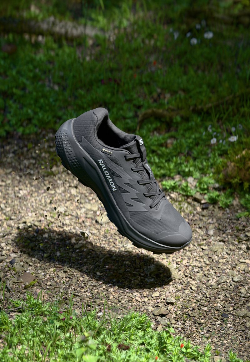 Black running shoe with textured upper, cushioned sole, and visible logo. Floating above a gravel path surrounded by green foliage.